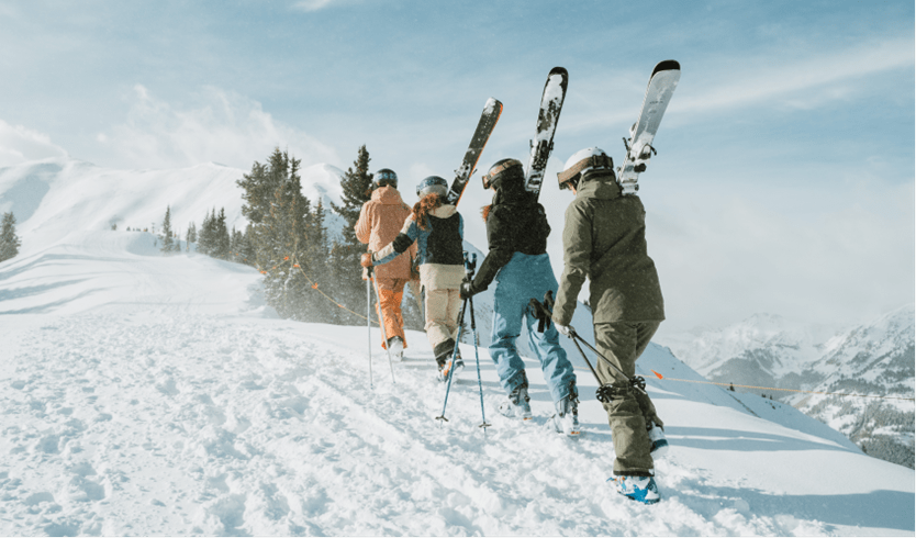 four people walk in single file up the highlands bowl with their skies on their shoulders, wind blows snow across the high alpine setting